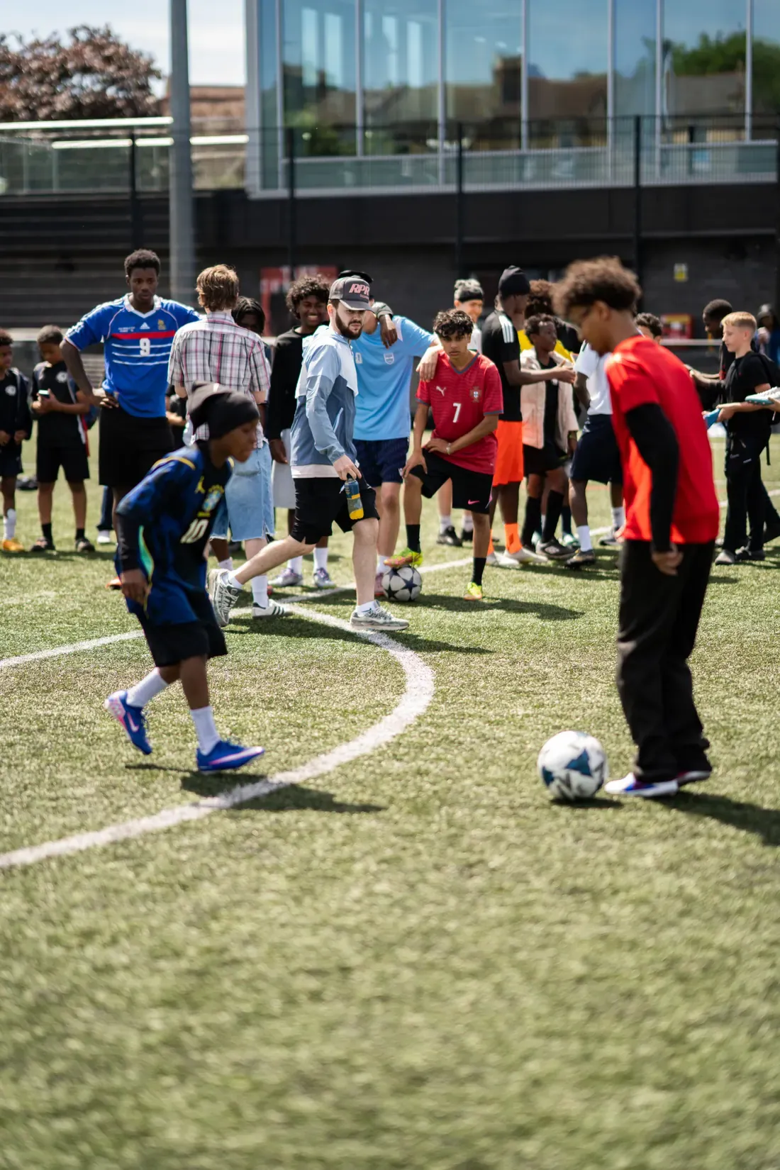 A grass pitch mid-shoot on a bright day, a boy running onto a ball with a crowd of players watching from the touchline