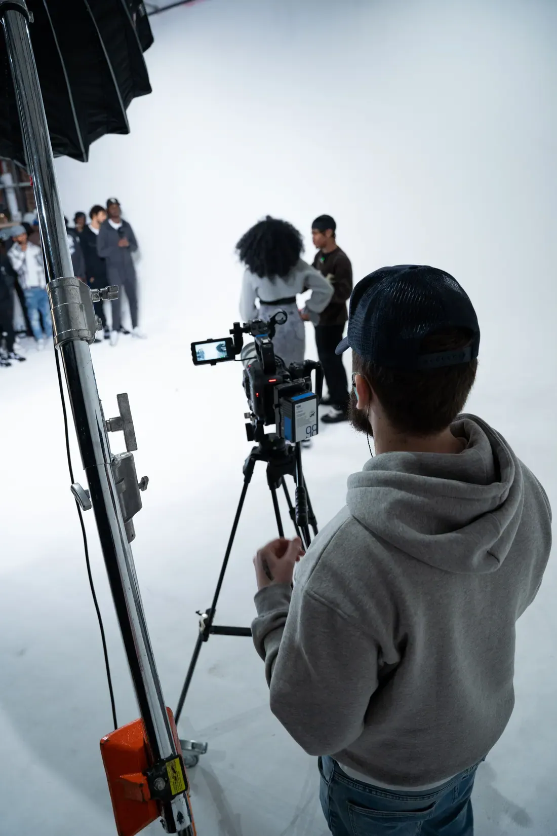Filming on a white cyc, seen from behind the operator: a camera on sticks with a phone clipped to it as a monitor, and contributors waiting at the far end