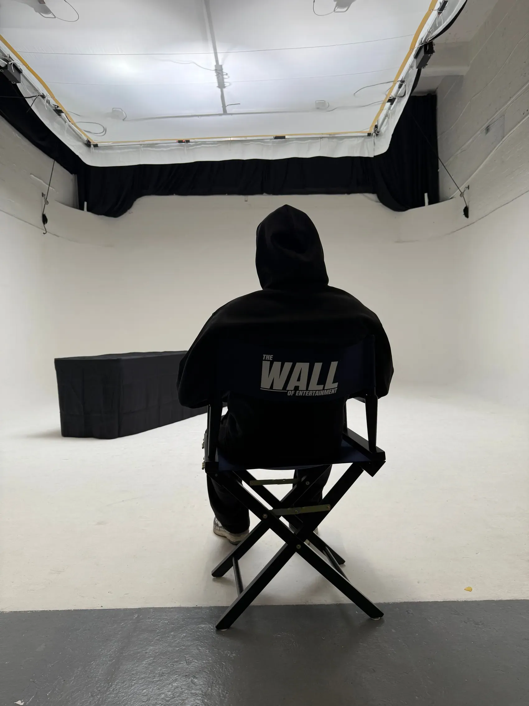 Someone sat with their back to camera in a black director's chair printed The Wall Of Entertainment, on a white cyc under a large overhead softbox