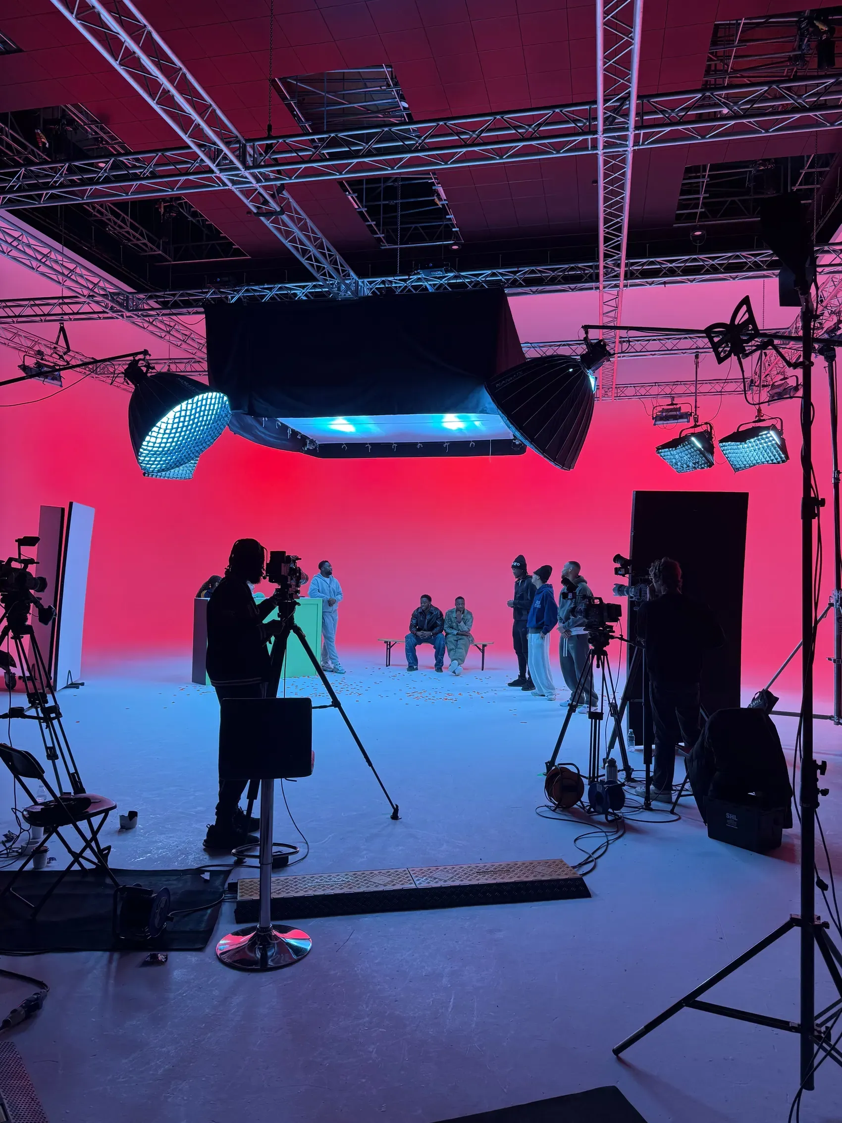 A studio floor mid-shoot: an infinity cyc lit red and blue, an operator on sticks in the foreground, contributors sat on apple boxes at the far end